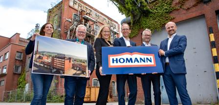 Sechs Personen mit zwei Schildern in den Händen stehen vor einem historischen Fabrikgebäude.