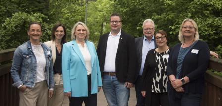 Sieben Personen stehen nebeneinander auf einem Weg mit Bäumen im Hintergrund.