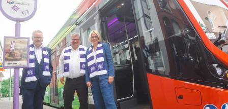 Drei Personen mit weiß-lila gestreiften Schals stehen vor einem Bus.