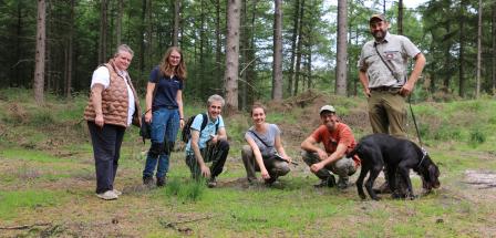Sechs Personen und ein schwarzer Hund stehen oder Hocken auf einer Waldwiese.
