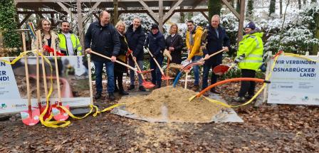 Eine Personengruppe mit Schaufeln steht hinter einem Sandhaufen.