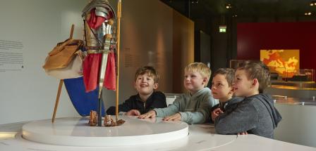 Vier Kinder stehen in einem Museumsraum und schauen eine römische Rüstung an.