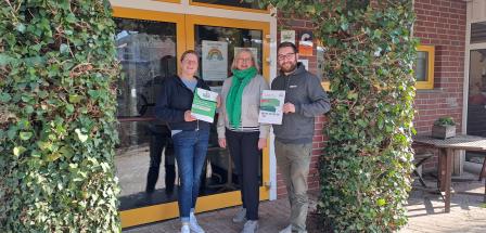 Drei Personen mit Urkunden in den Händen stehen im begrünten Eingangsbereich eines Gebäudes.