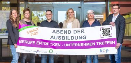 Sechs Personen mit einem Werbebanner in den Händen stehen vor einem Gebäudeeingang.