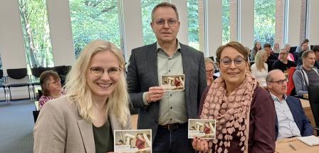 Drei Personen in einem Versammlungssaal halten Flyer in die Kamera.