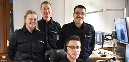 Vier Personen in Uniform stehen und sitzen in einer Rettungsleitstelle.