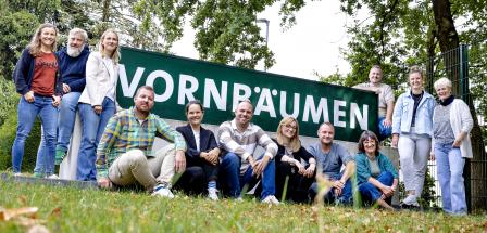 Eine Personengruppe steht, hockt und sitzt vor einem Zaun.