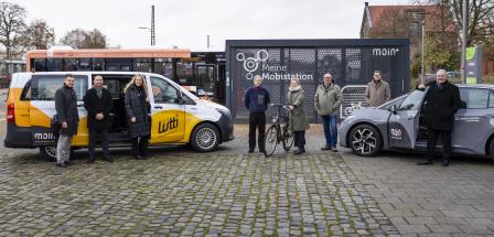 Eine Personengruppe steht vor einem Kleinbus und einem PKW.