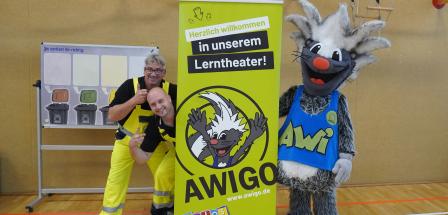 Zwei Personen und ein Maskottchen stehen neben einem Roll Up Poster.