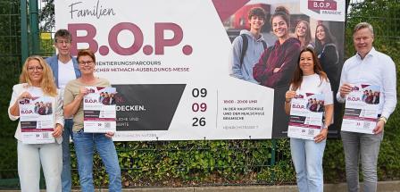Fünf Personen stehen vor einem Zaun, an dem ein Plakat befestigt ist.