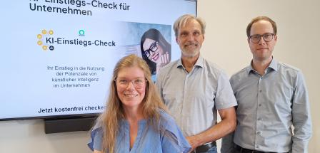 Drei Personen stehen vor einem großen Bildschirm.