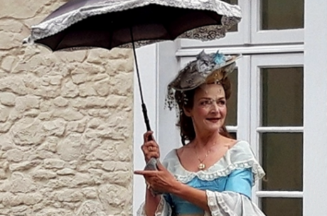 Eine Person im historischen Kostüm mit Schirm steht vor einem Haus.