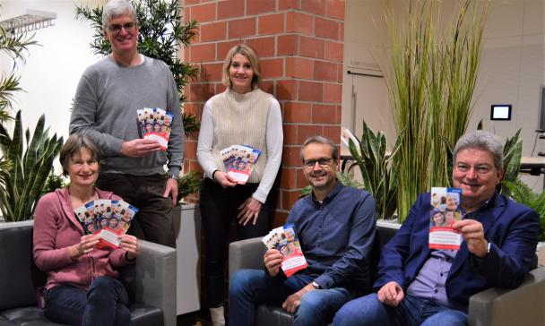 Fünf Personen sitzen in einer grauen Sitzgruppe, zwei stehen und halten Flyer in die Kamera.