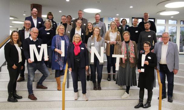 Eine Personengruppe steht in einem Gebäude auf einer Treppe. Einige halten Buchstaben in den Händen, die zusammen das Wort MINT ergeben.
