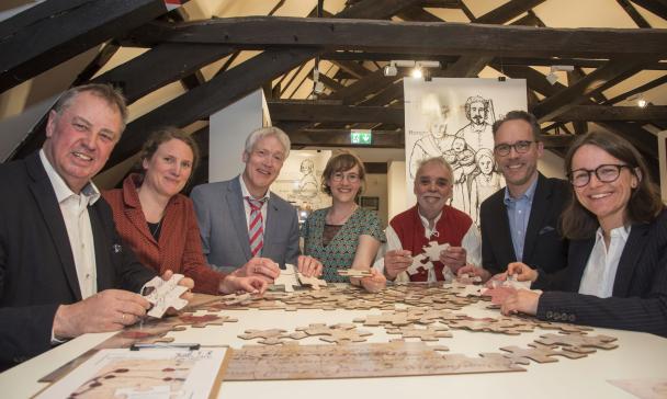 Sieben Personen stehen um einen Tisch herum, auf dem ein Puzzle liegt.