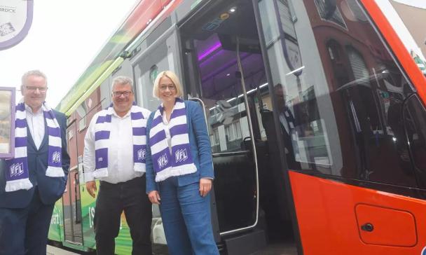 Drei Personen mit weiß-lila gestreiften Schals stehen vor einem Bus.
