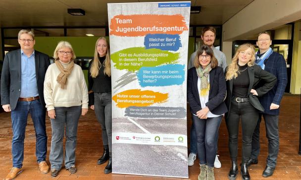 Sieben Personen stehen im Eingangsbereich eines Gebäudes links und rechts neben einem Rollup-Poster.