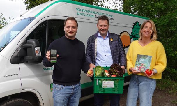Drei Personen stehen vor einem Lieferwagen und halten Lebensmittel in die Kamera.