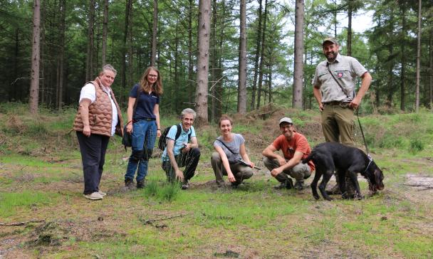 Sechs Personen und ein schwarzer Hund stehen oder Hocken auf einer Waldwiese.