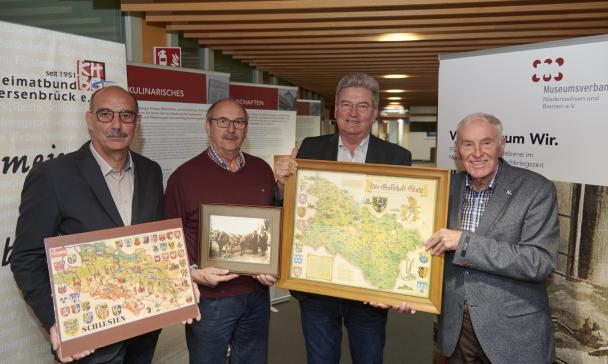 Vier Personen stehen vor den Postern einer Ausstellung und halten historische Karten in den Händen.