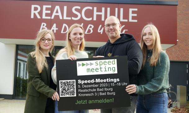 Vier Personen mit einem Schild in den Händen stehen vor einem Gebäudeeingang.