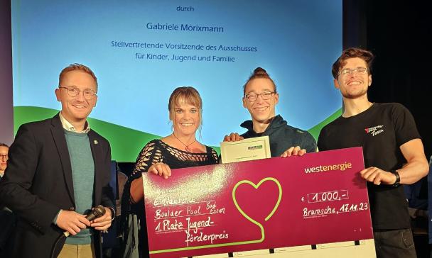Vier Personen stehen mit einem symbolischen Scheck auf einer Bühne vor einer Leinwand.
