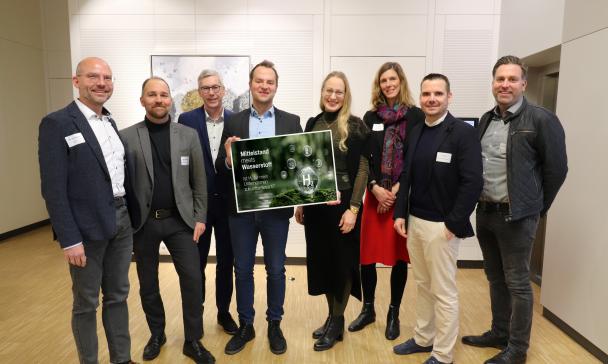 Eine Personengruppe steht in einem Foyer und hält eine Poster in die Kamera.