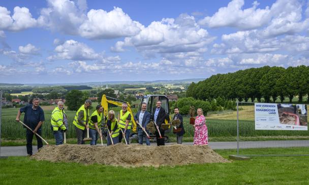 Eine Personengruppe mit Schaufeln in den Händen steht hinter einem Sandhaufen.