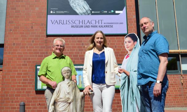 Drei Personen stehen vor einem großen Plakat an einer Hauswand.