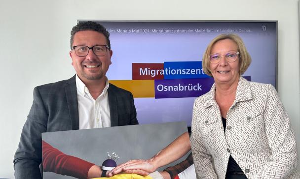 Zwei Personen mit einem großen Foto in den Händen stehen vor einem Display.