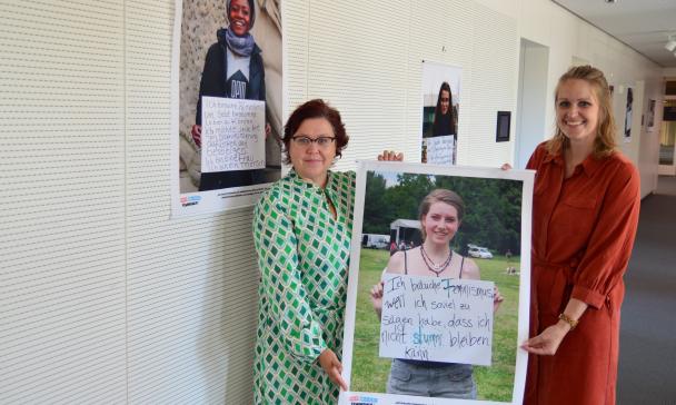 Zwei Personen mit einem Poster in den Händen stehen in einem Gebäudeflur.
