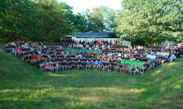 Eine große Personengruppe sitzt wie in einem Amphitheater auf einer Wiese.