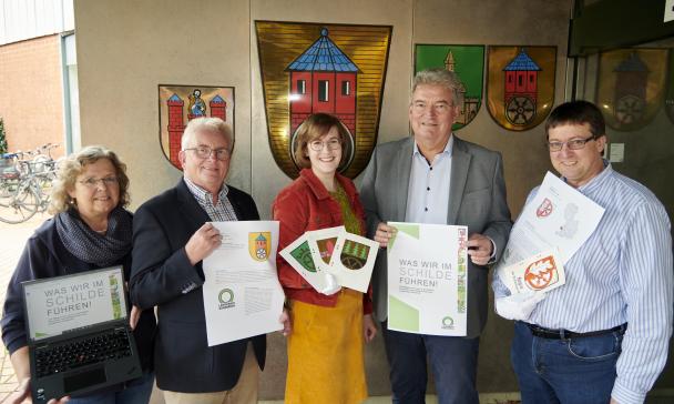 Fünf Personen stehen vor einer mit Wappen geschmückten Wand und präsentieren eine Broschüre.