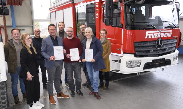 Eine Personengruppe steht neben einem Feuerwehrauto in einer Werkhalle.