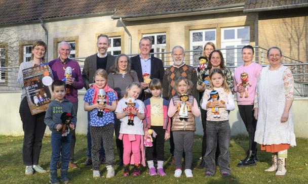 Eine Personengruppe mit Playmobilfiguren in den Händen steht vor einem Gebäude.