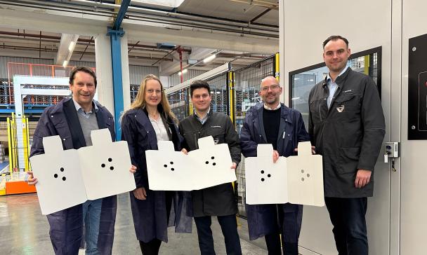 Fünf Personen mit Verpackungsmaterial in den Händen stehen in einer Werkhalle.