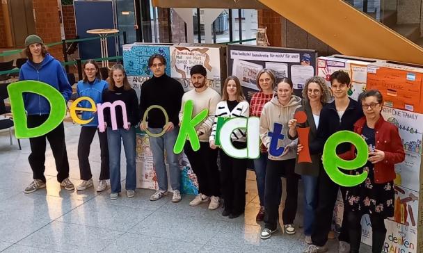 Eine Personengruppe steht in einem Foyer und hält Buchstaben in den Händen, die das Wort Demokratie bilden.