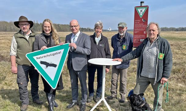 Eine Personengruppe steht am Rande eines Heidegebietes und hält ein Naturschutzschild in die Kamera.
