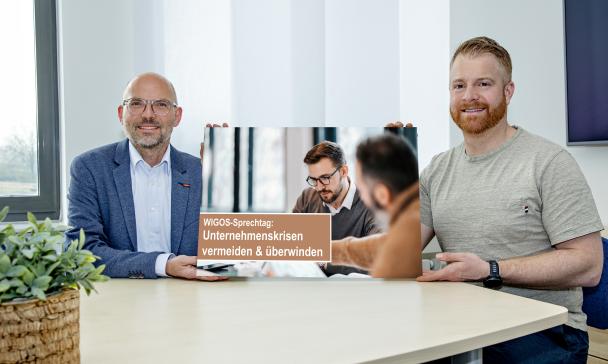 Zwei Personen sitzen an einem Tisch und halten eine Grafik in die Kamera.