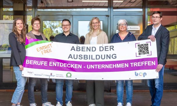 Sechs Personen mit einem Werbebanner in den Händen stehen vor einem Gebäudeeingang.