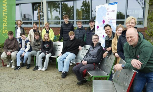 Eine Personengruppe sitzt auf Bänken vor einem Gebäude.