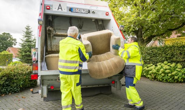 Zwei Personen entsorgen einen Sessel in einem Müllfahrzeug.