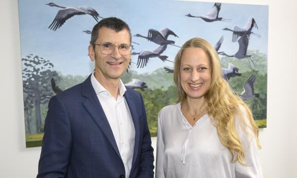 Zwei Personen stehen nebeneinander im Hintergrund ein Bild mit Vögeln.