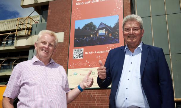 Zwei Personen stehen vor einem großen Plakat an einer Fassade.