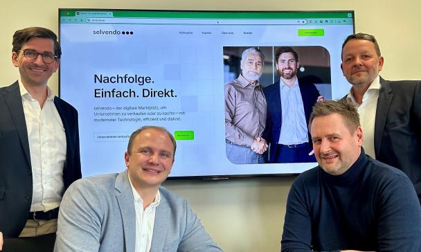 Vier Personen sitzen vor und stehen neben einem großen Bildschirm.