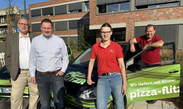 Vier Personen stehen neben dem Auto eines Pizzalieferdienstes.