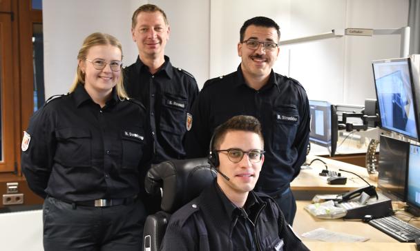 Vier Personen in Uniform stehen und sitzen in einer Rettungsleitstelle.
