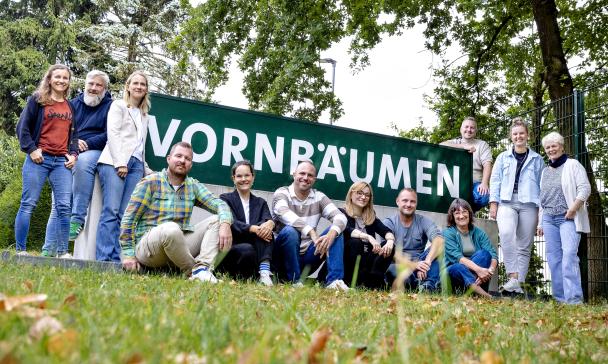 Eine Personengruppe steht, hockt und sitzt vor einem Zaun.