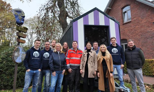 Personengruppe vor einem kleinen Holzhaus