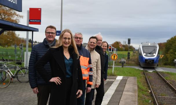 Eine Personengruppe steht auf einem Bahnsteig. Im Hintergrund nähert sich ein Zug.